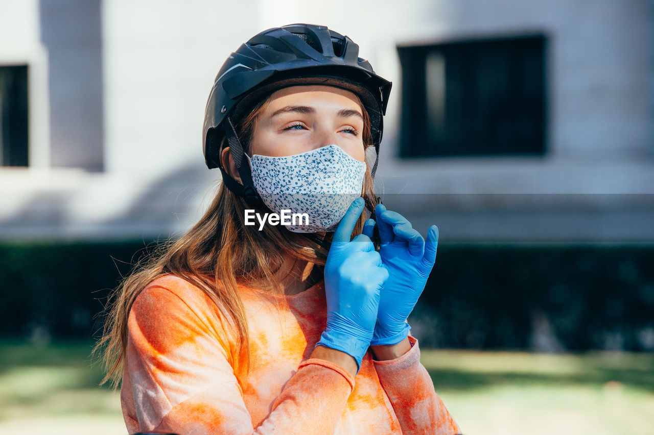 Female cyclist in latex gloves and fabric mask looking away and fastening hardhat on blurred background of street of madrid, spain
