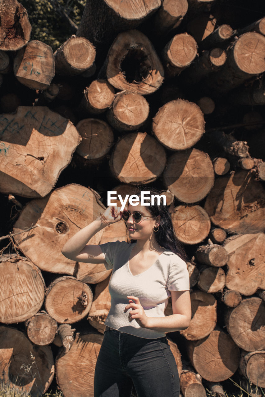 Woman holding sunglasses while standing against stack of firewood