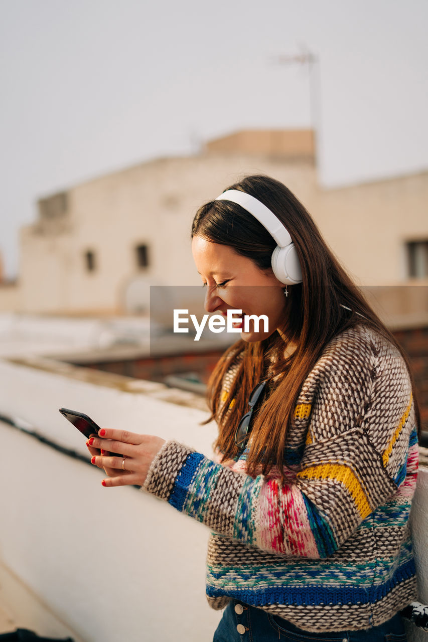 Smiling woman wearing headphones and using phone on building terrace