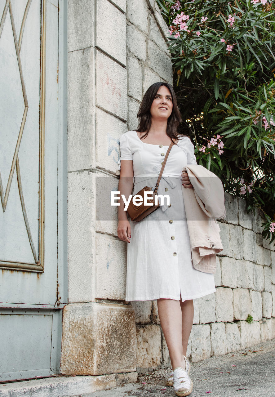 Full length image of beautiful woman in white summer dress leaning on stone wall.