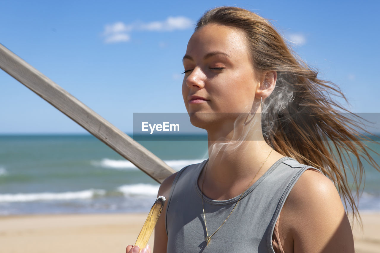 Beautiful young woman meditating by the beach with palo santo