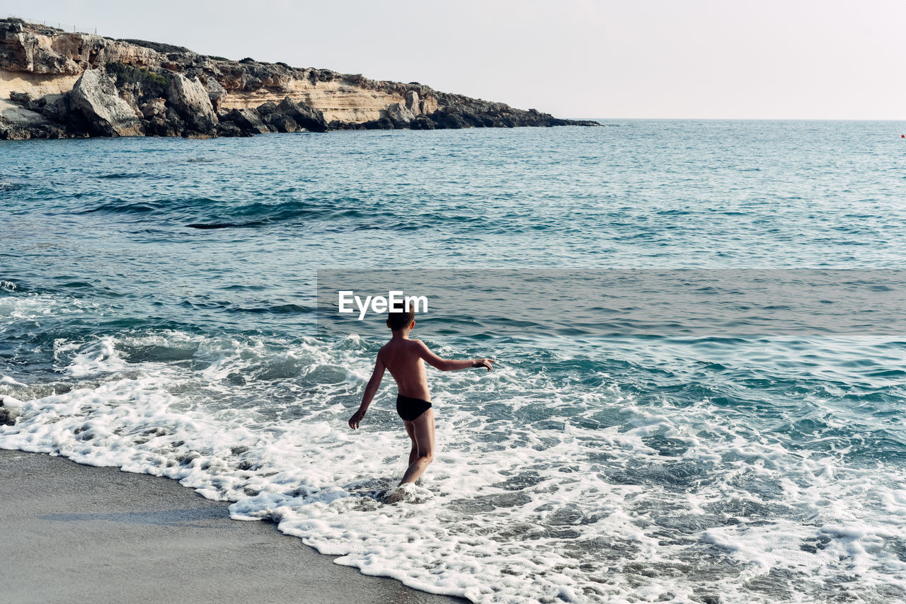 Shirtless boy standing in sea against clear sky