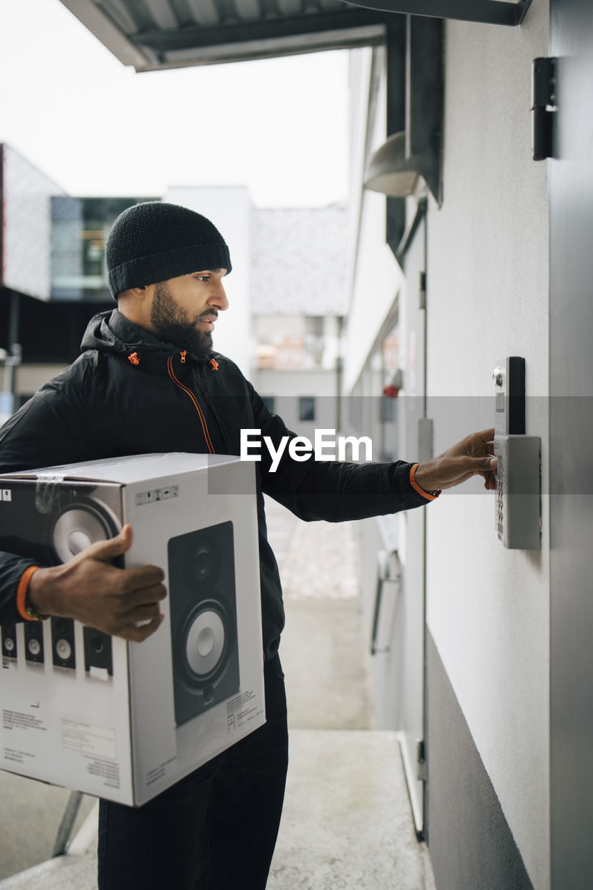 Serious male worker ringing intercom while carrying box during delivery