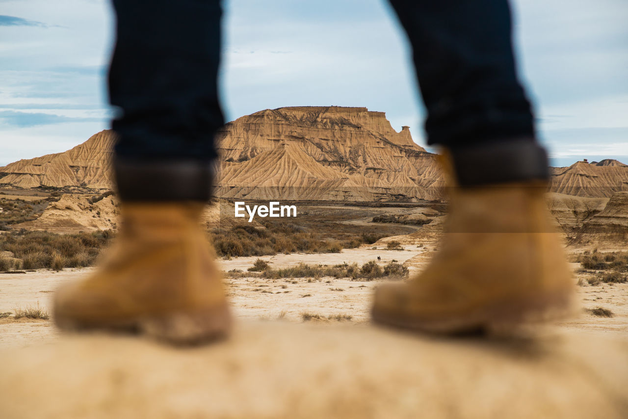 Legs on faceless traveler in brown boots and blue jeans standing on dirty sandy road with mountain and sky on blurred background in bardenas reales, navarre, spain