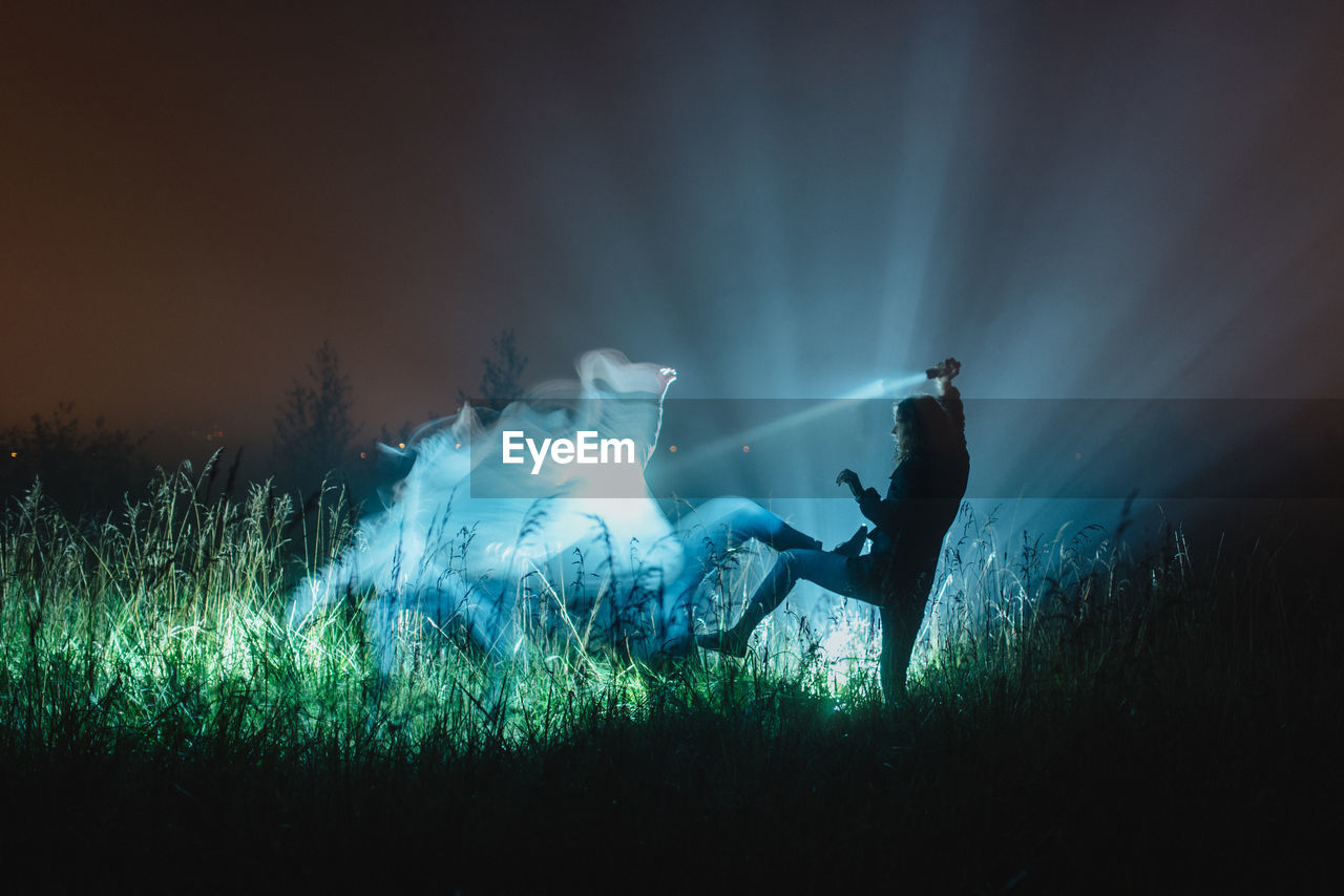 Young woman holding flashlight over digitally composite man on field at night