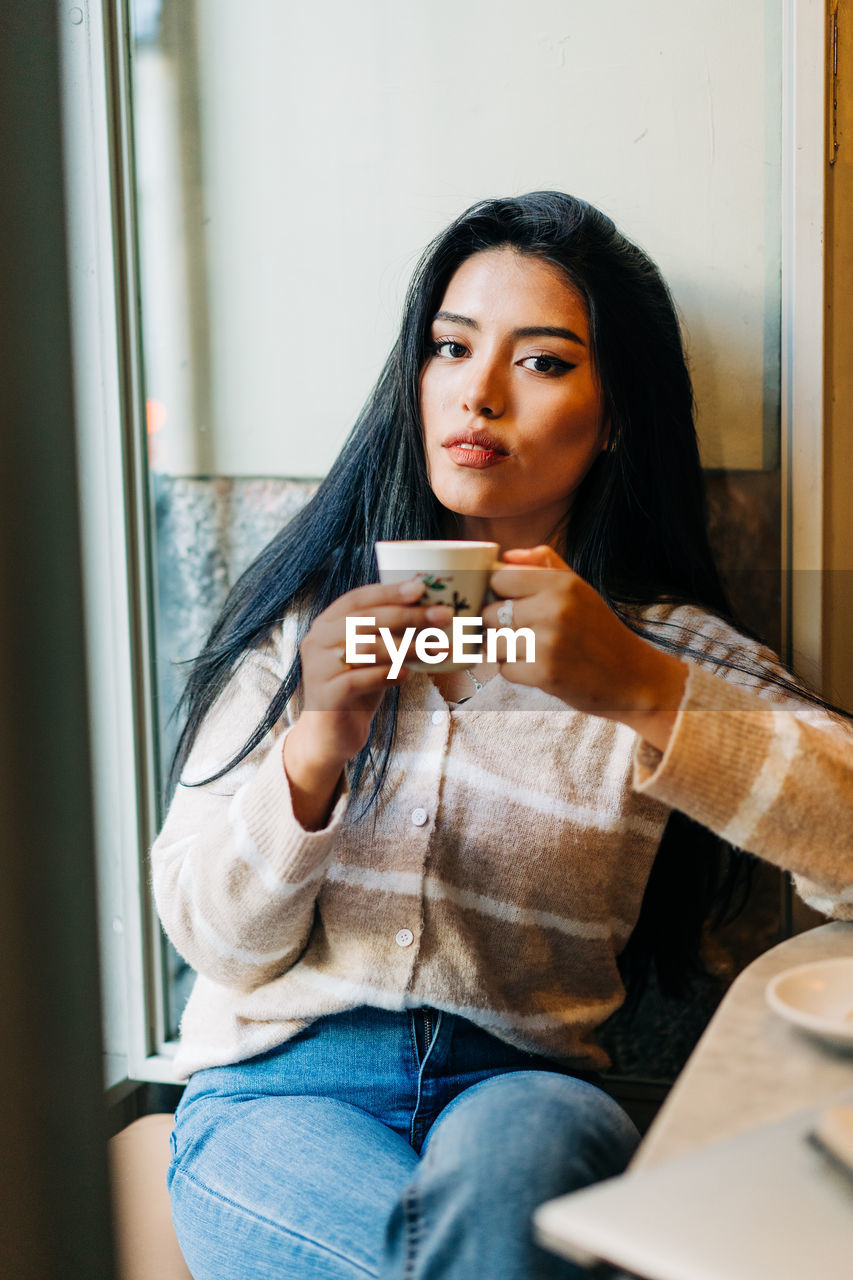 Ethnic female with makeup drinking hot beverage from cup in coffee house