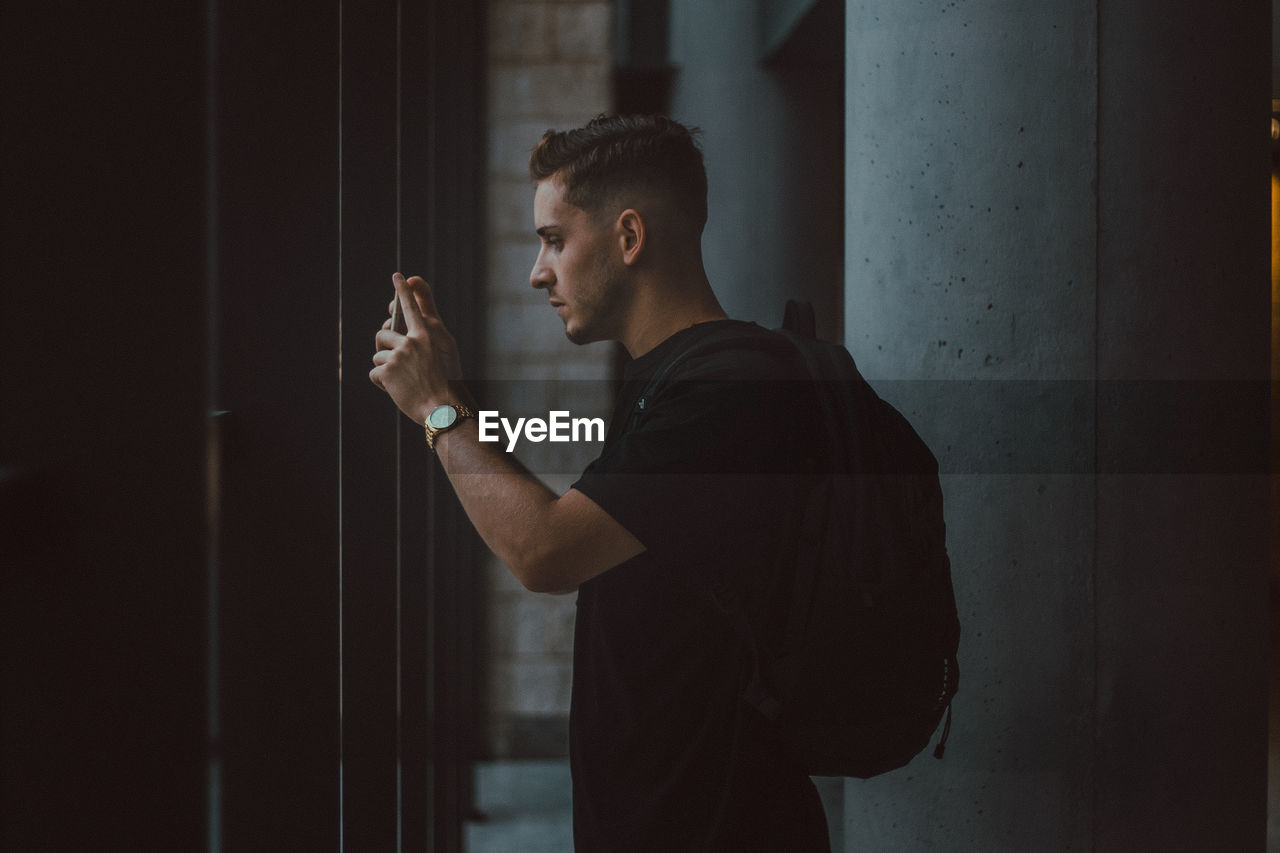 Side view of young man photographing while standing against wall