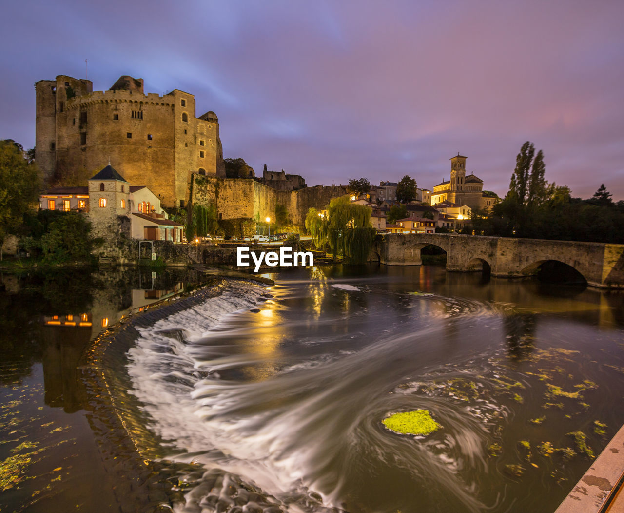 Illuminated buildings by river against sky at dusk