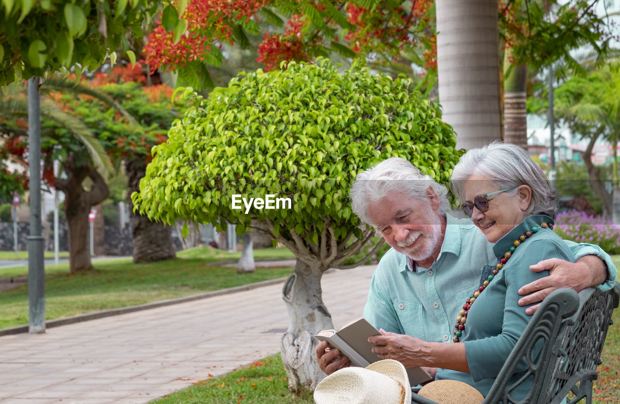 Smiling senior couple sitting on bench reading book at park
