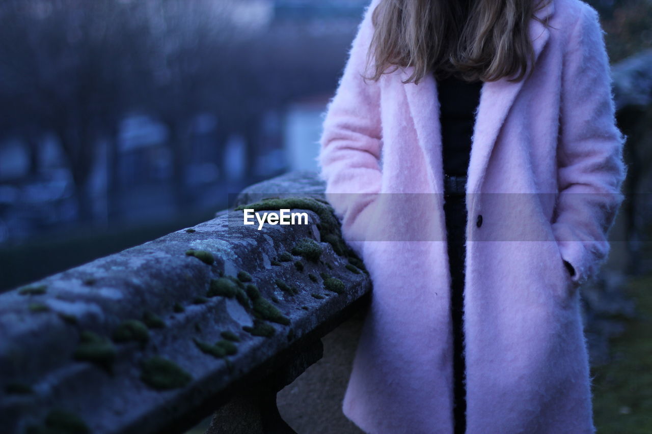 Midsection of woman wearing pink long coat standing by railing at dusk