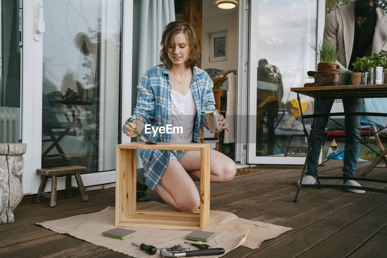 Young woman applying paint on wooden furniture while kneeling on porch