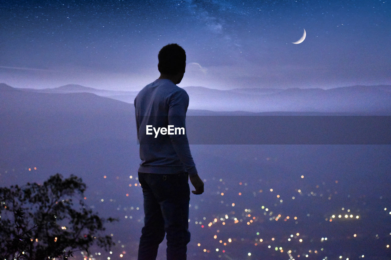 REAR VIEW OF MAN LOOKING AT MOON AT DUSK