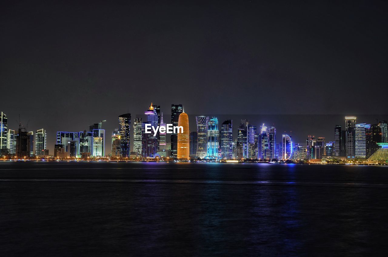 Illuminated buildings by sea against sky at night