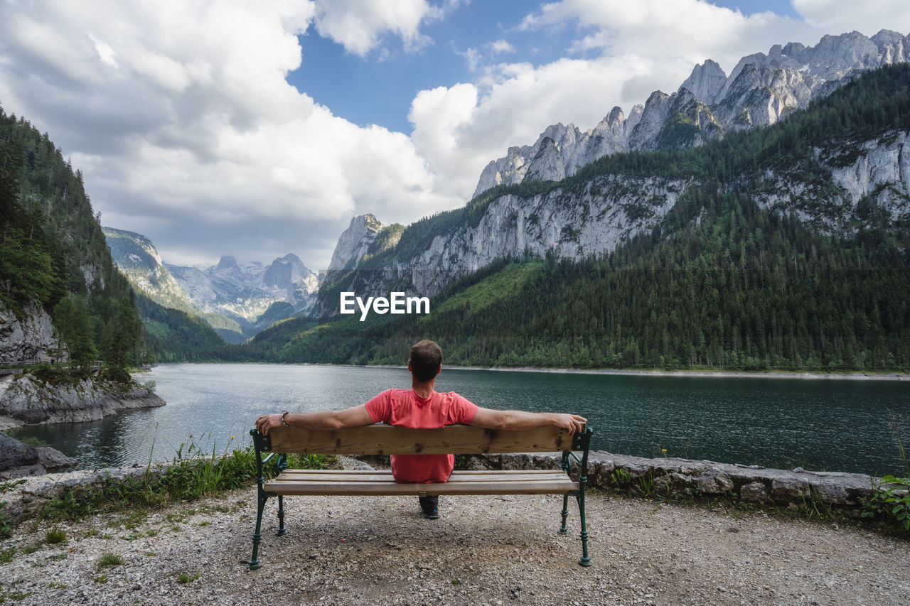 WOMAN SITTING ON BENCH BY LAKE AGAINST MOUNTAINS