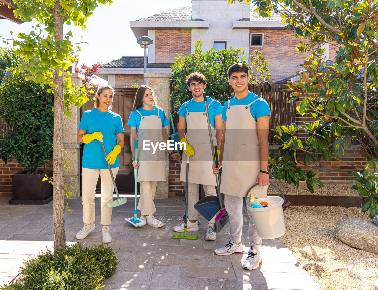 Full body of happy young male and female coworkers in uniforms and gloves smiling while standing near modern brick residential house with brooms and buckets after cleaning service