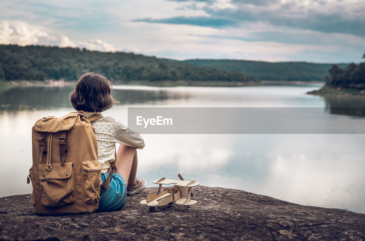 REAR VIEW OF MAN SITTING ON ROCK LOOKING AT LAKE