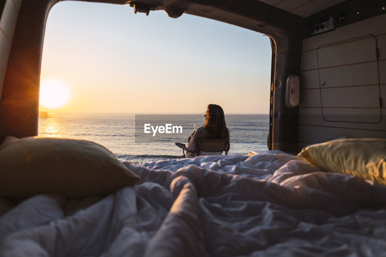 Woman admiring sunset view while while sitting on chair by camper van at beach