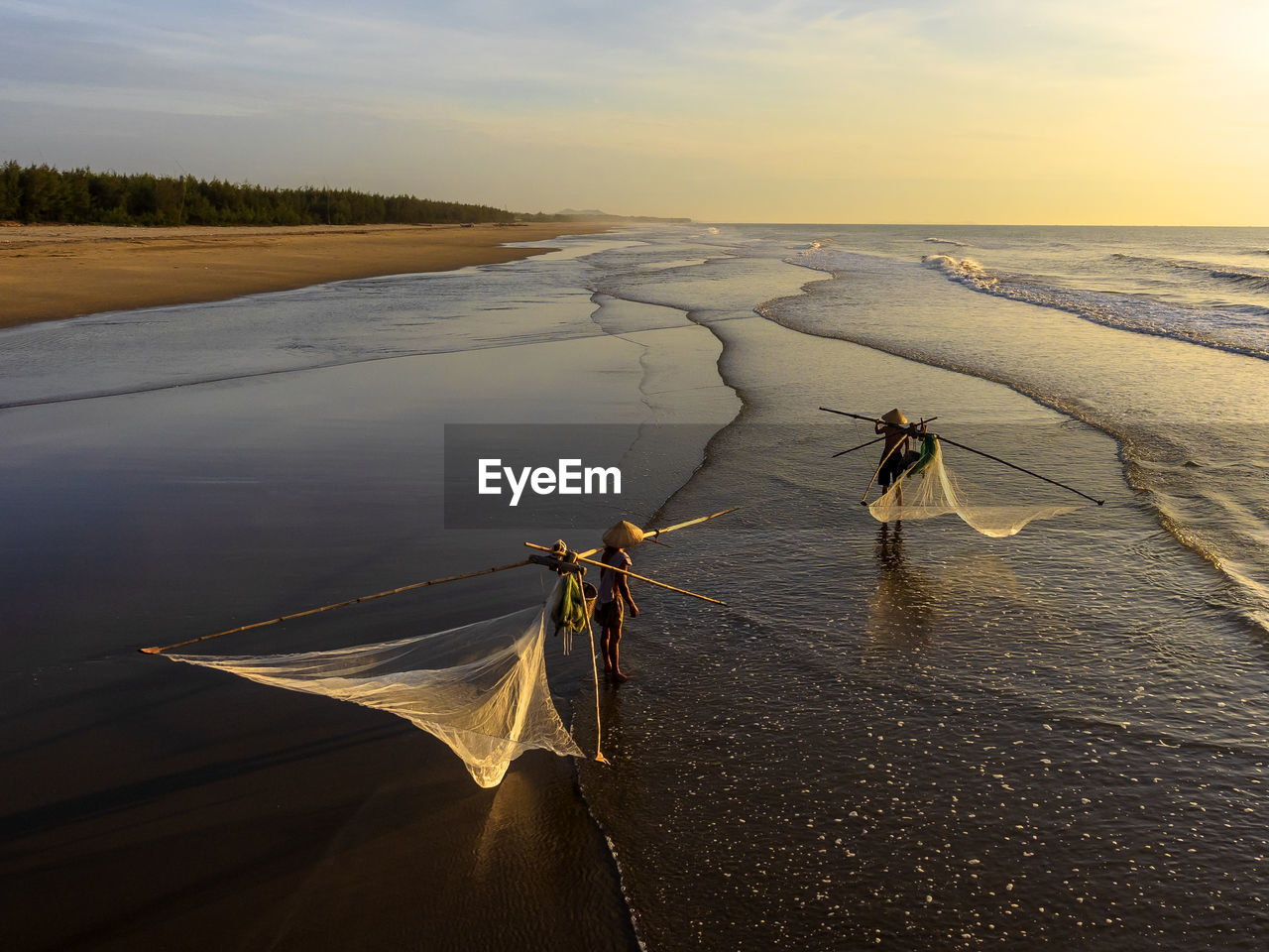 Fishermen with fishing nets standing at beach against sky during sunset