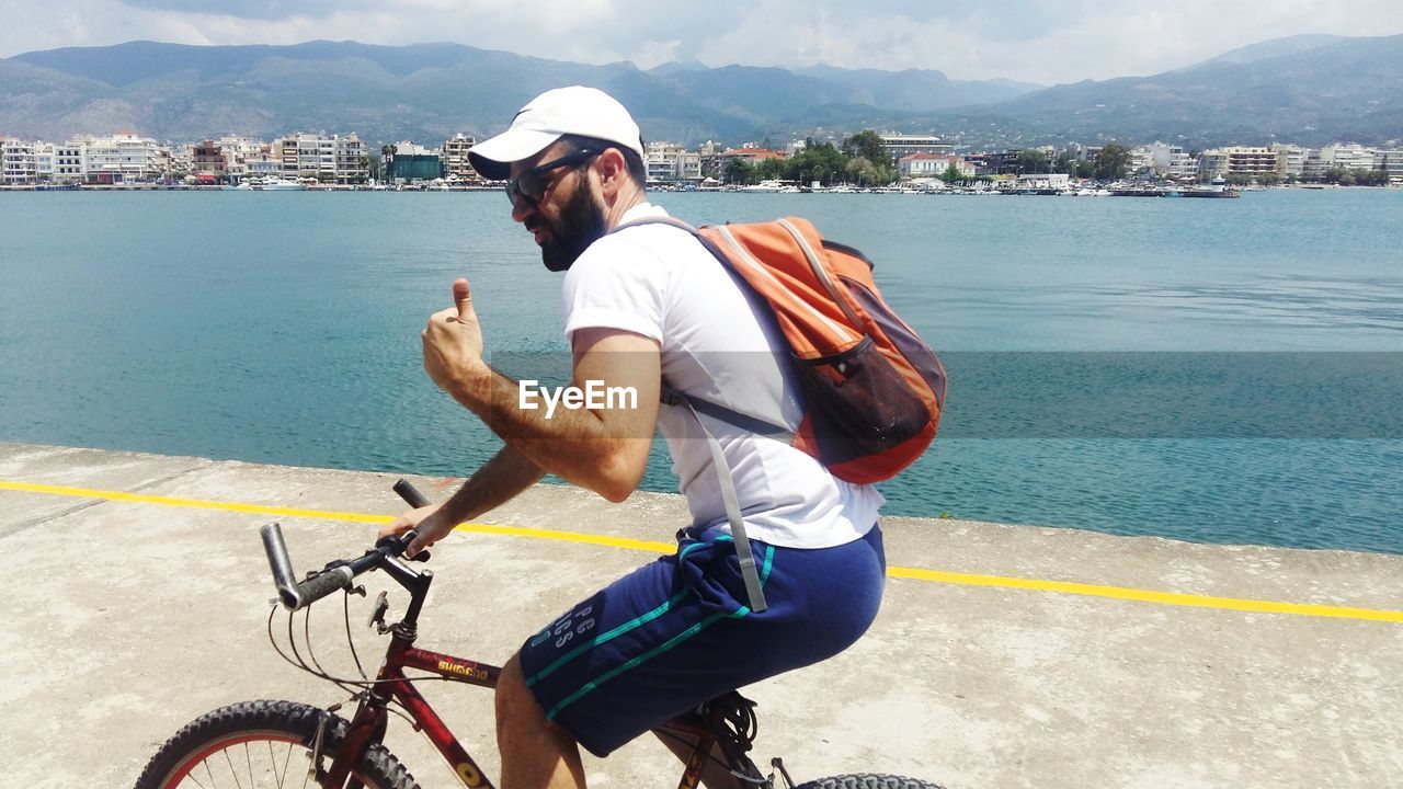 Man showing thumbs up sign while riding bicycle on road by lake in town