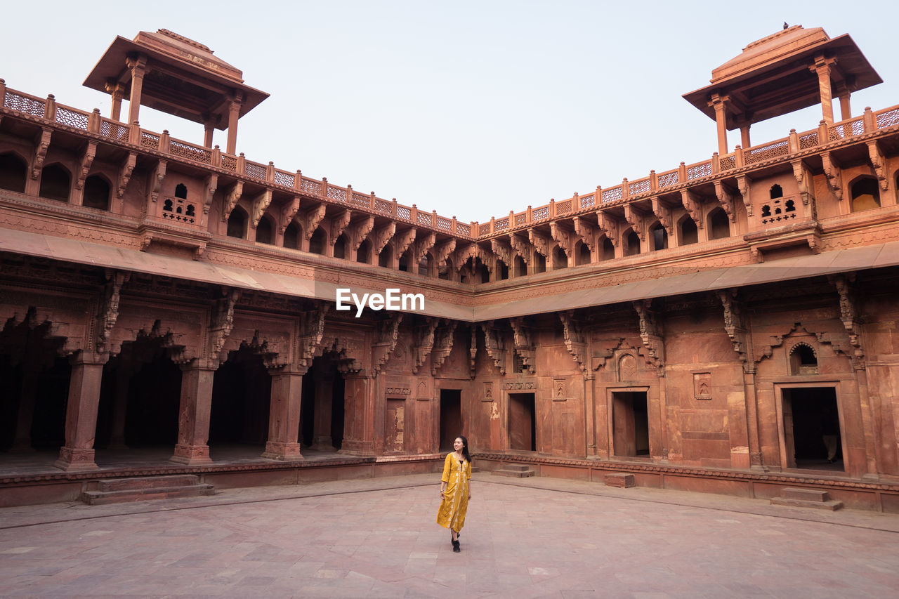 Ethnic asian woman looking away in yellow dress standing in aged arched agra fort in uttar pradesh, india