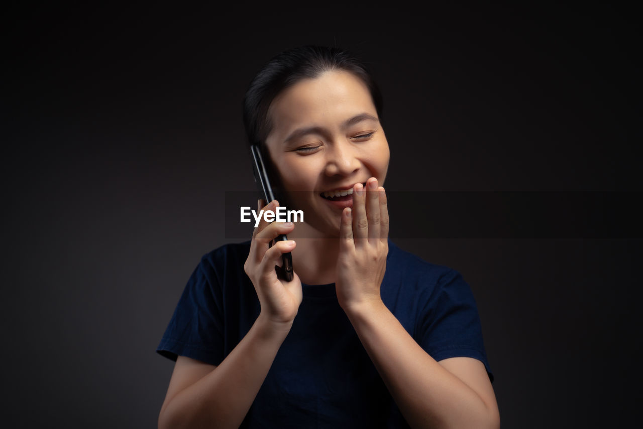 SMILING YOUNG WOMAN USING PHONE AGAINST BLACK BACKGROUND