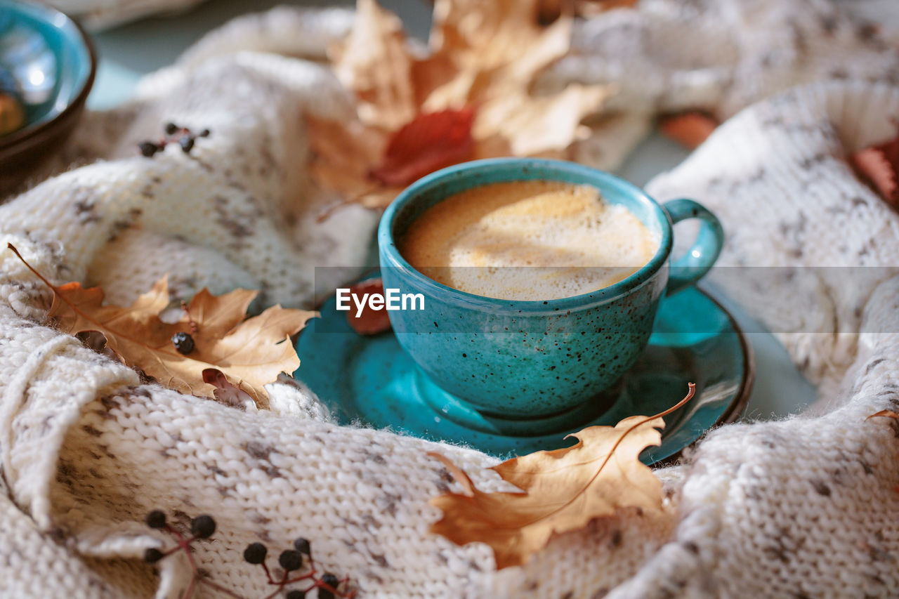 White knitted scarf, blue cup of coffee and dry yellow leaves