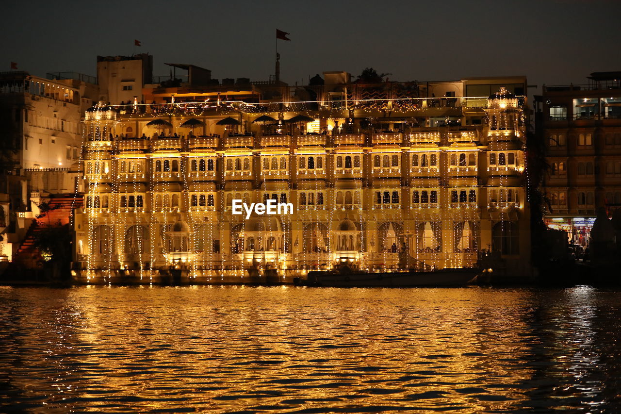 illuminated buildings by river against sky at night