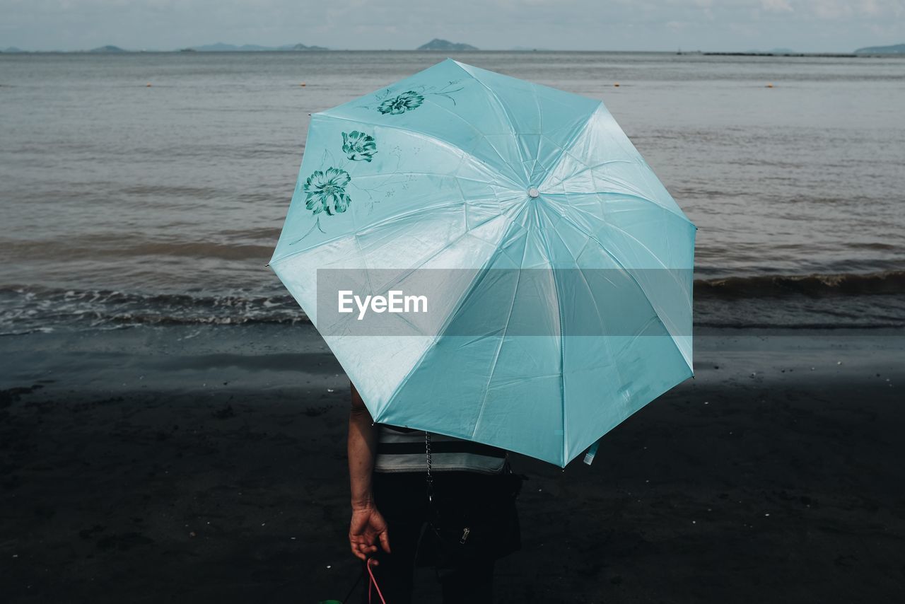Rear view of man with umbrella standing on beach during monsoon