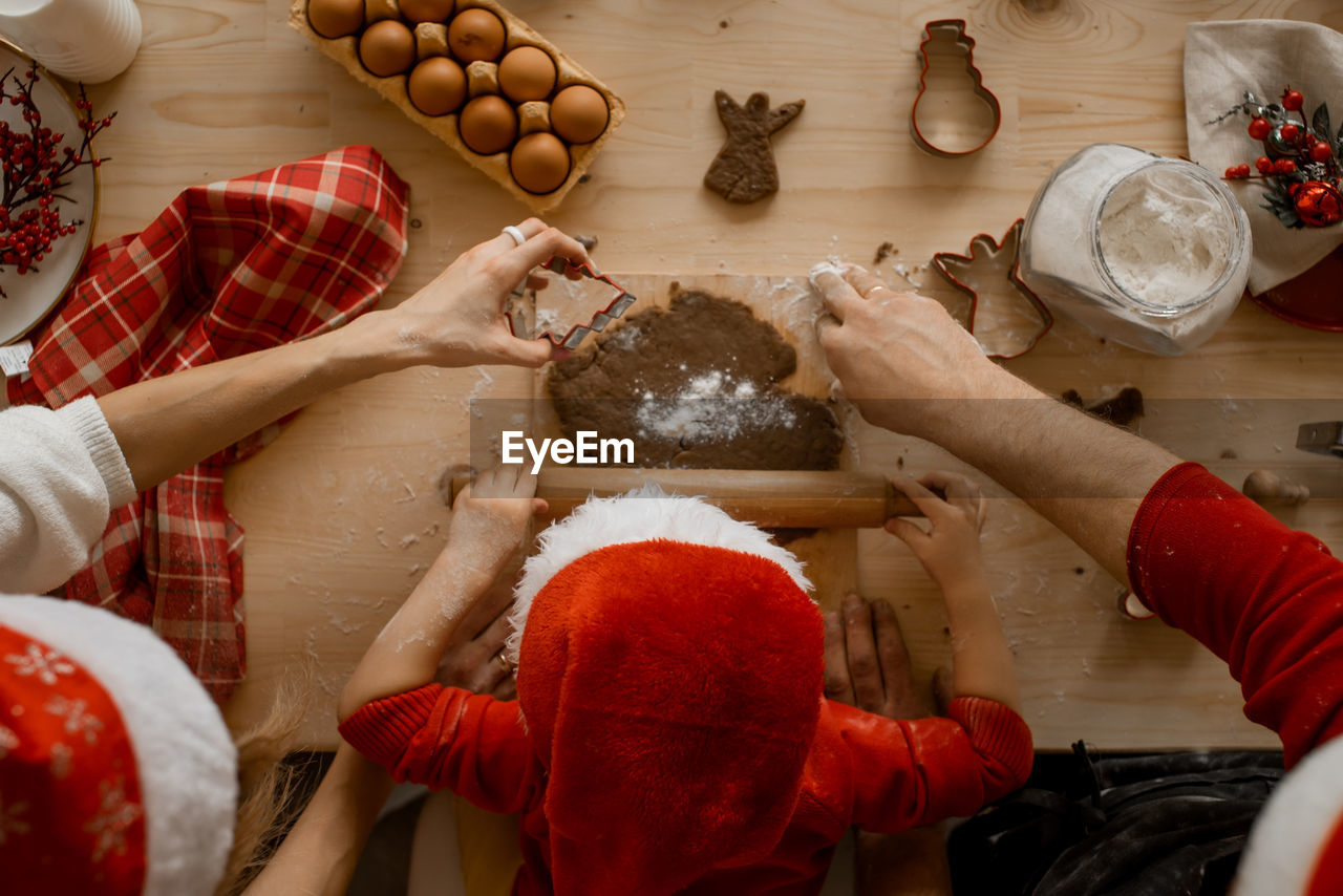A top view of the hands of mather, father and child in a santa hat, rolling dough with a rock