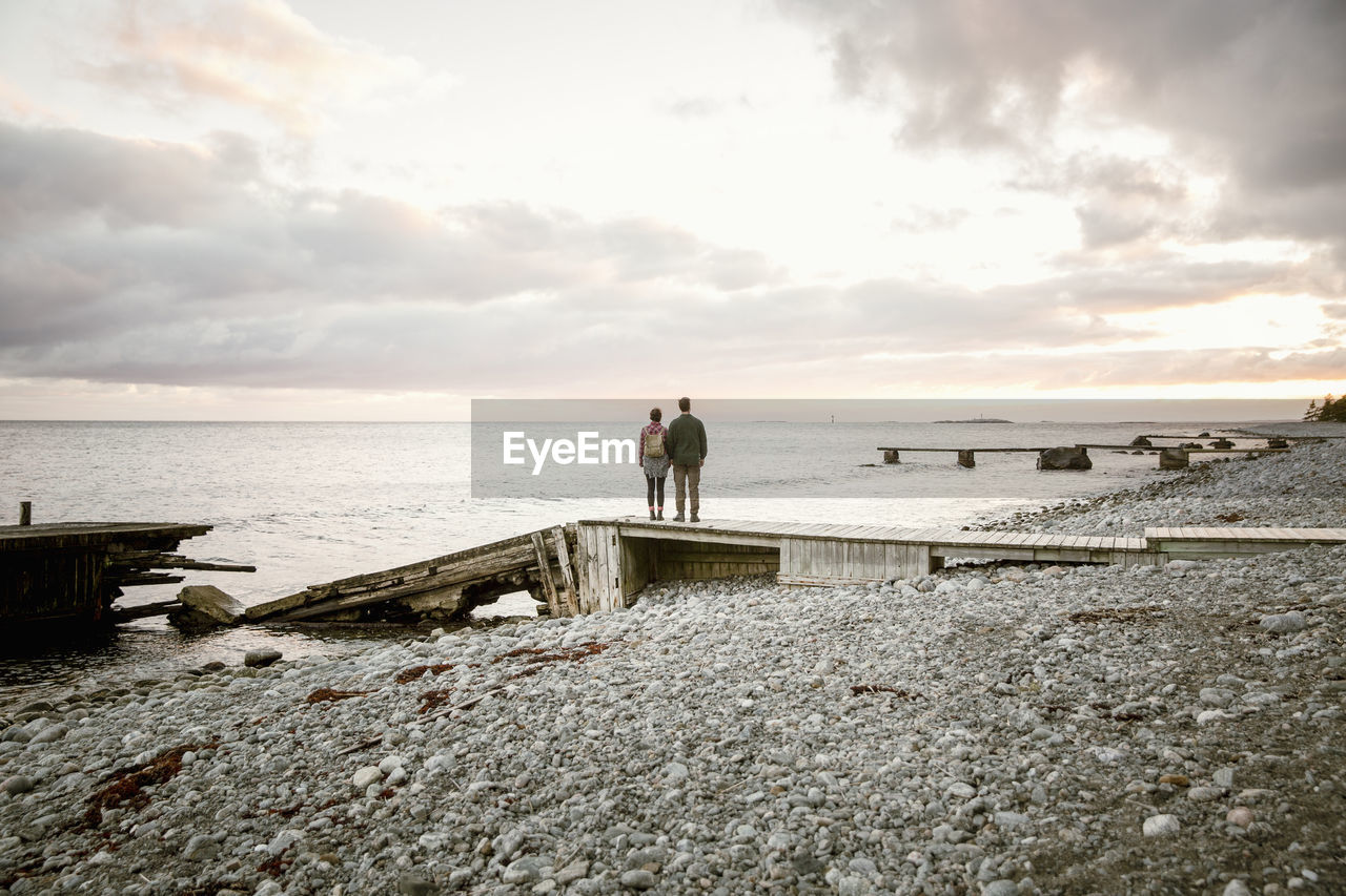 Rear view of couple standing on jetty at beach against sky during sunset