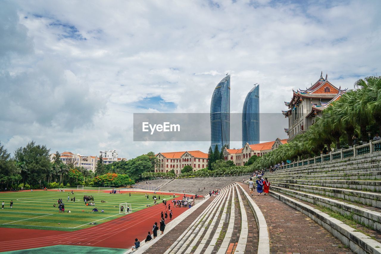 People at soccer stadium by modern buildings against cloudy sky