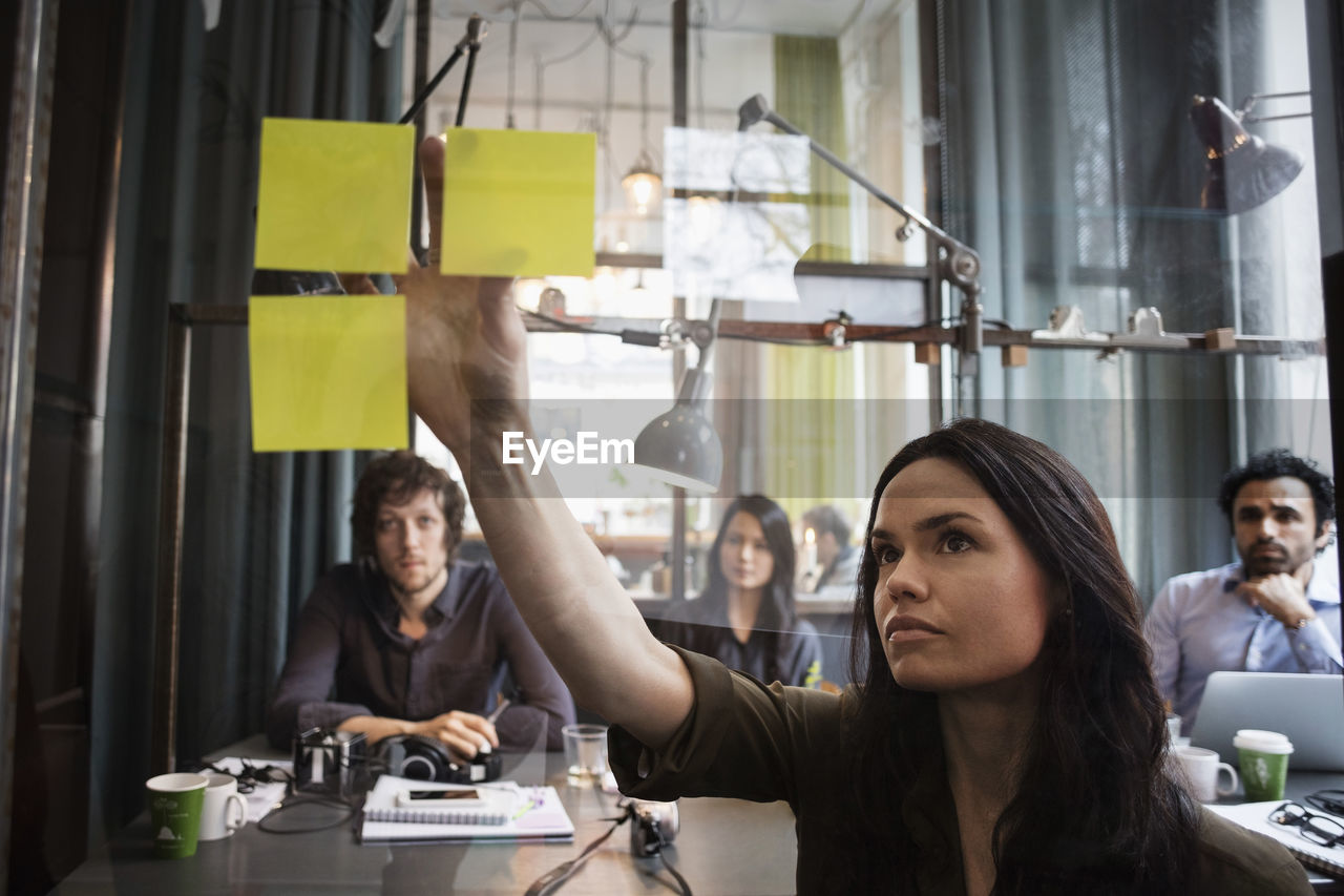 Creative business people looking at female colleague sticking adhesive note on window seen through glass