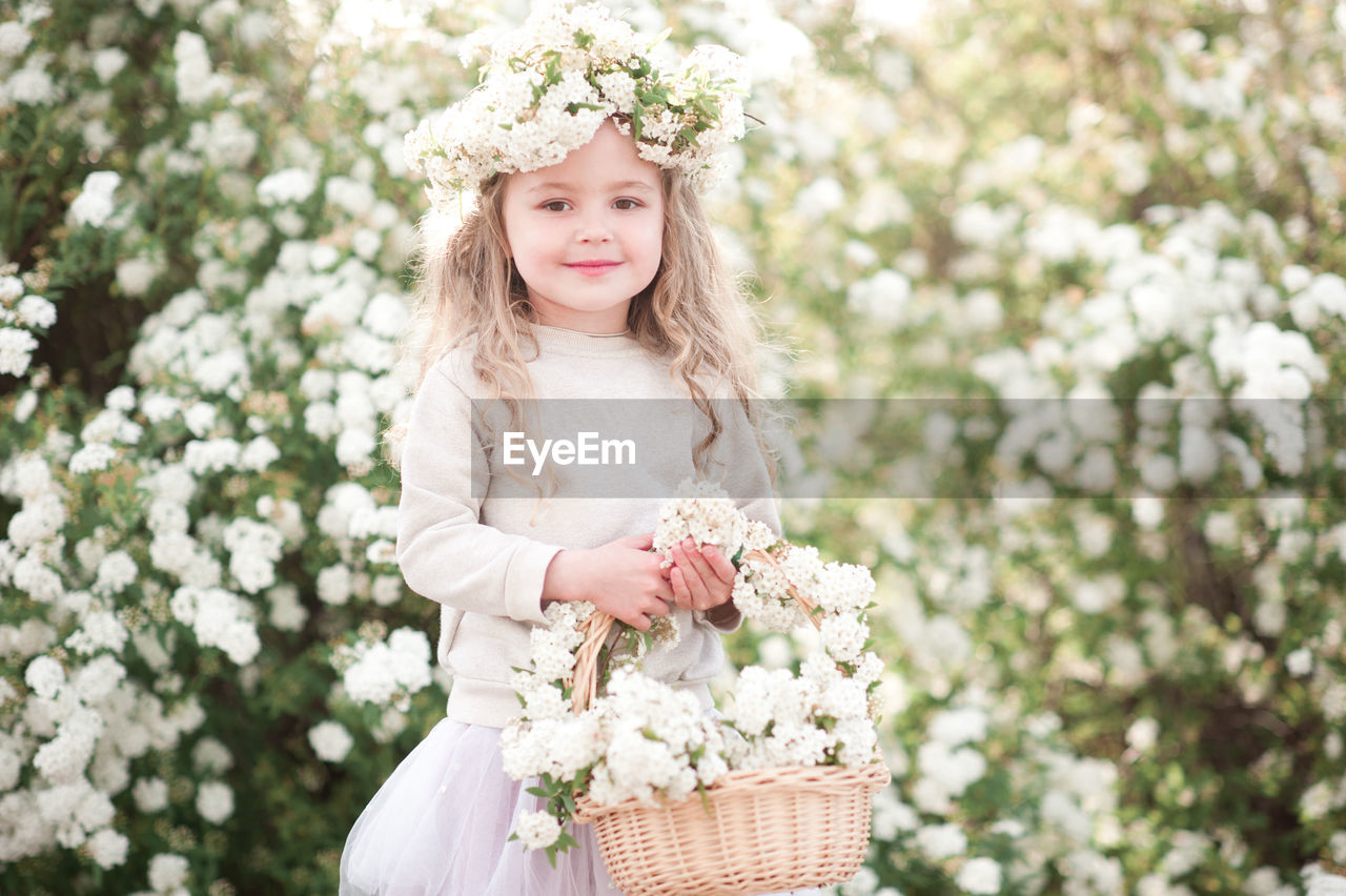 flower, plant, flowering plant, women, one person, nature, spring, child, bride, clothing, portrait, childhood, smiling, long hair, happiness, beauty in nature, dress, freshness, hairstyle, female, blond hair, front view, fashion, emotion, white, flower arrangement, adult, looking at camera, basket, wreath, wedding dress, innocence, pink, bouquet, outdoors, three quarter length, springtime, day, celebration, portrait photography, lifestyles, young adult, standing, cute, summer, blossom, looking, lilac, laurel wreath, tree, person, crown, event, holding