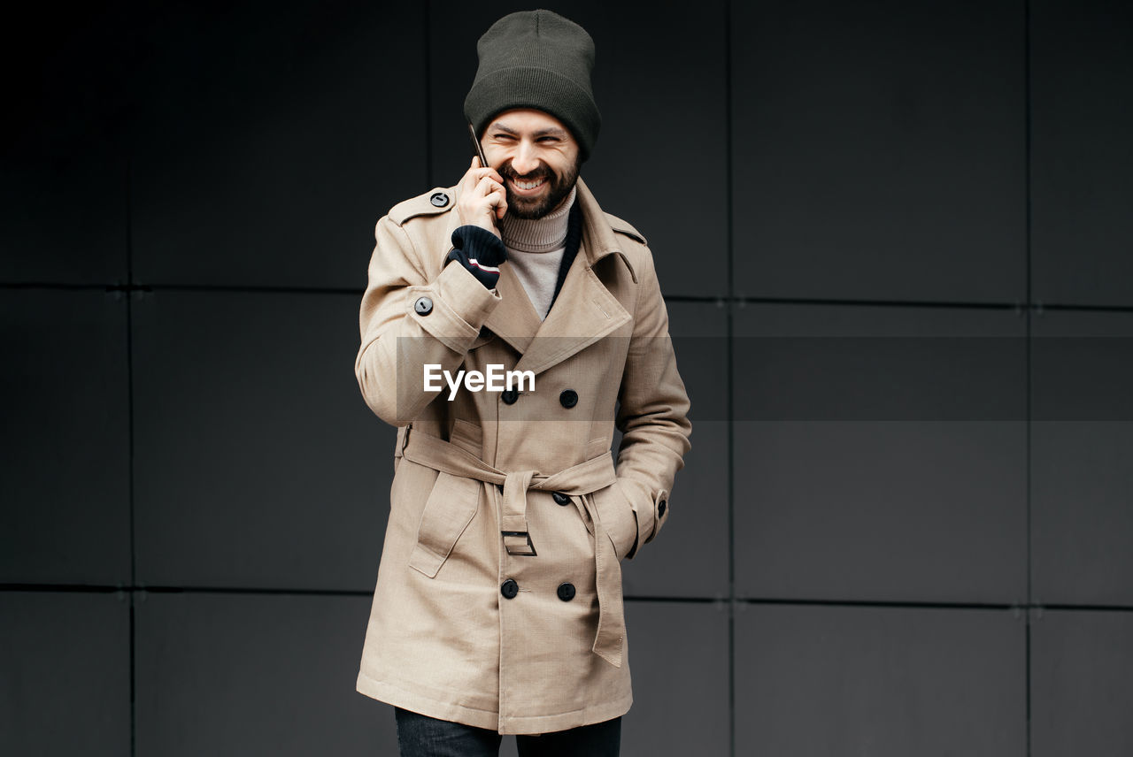 Portrait of smiling man talking on phone while standing against wall