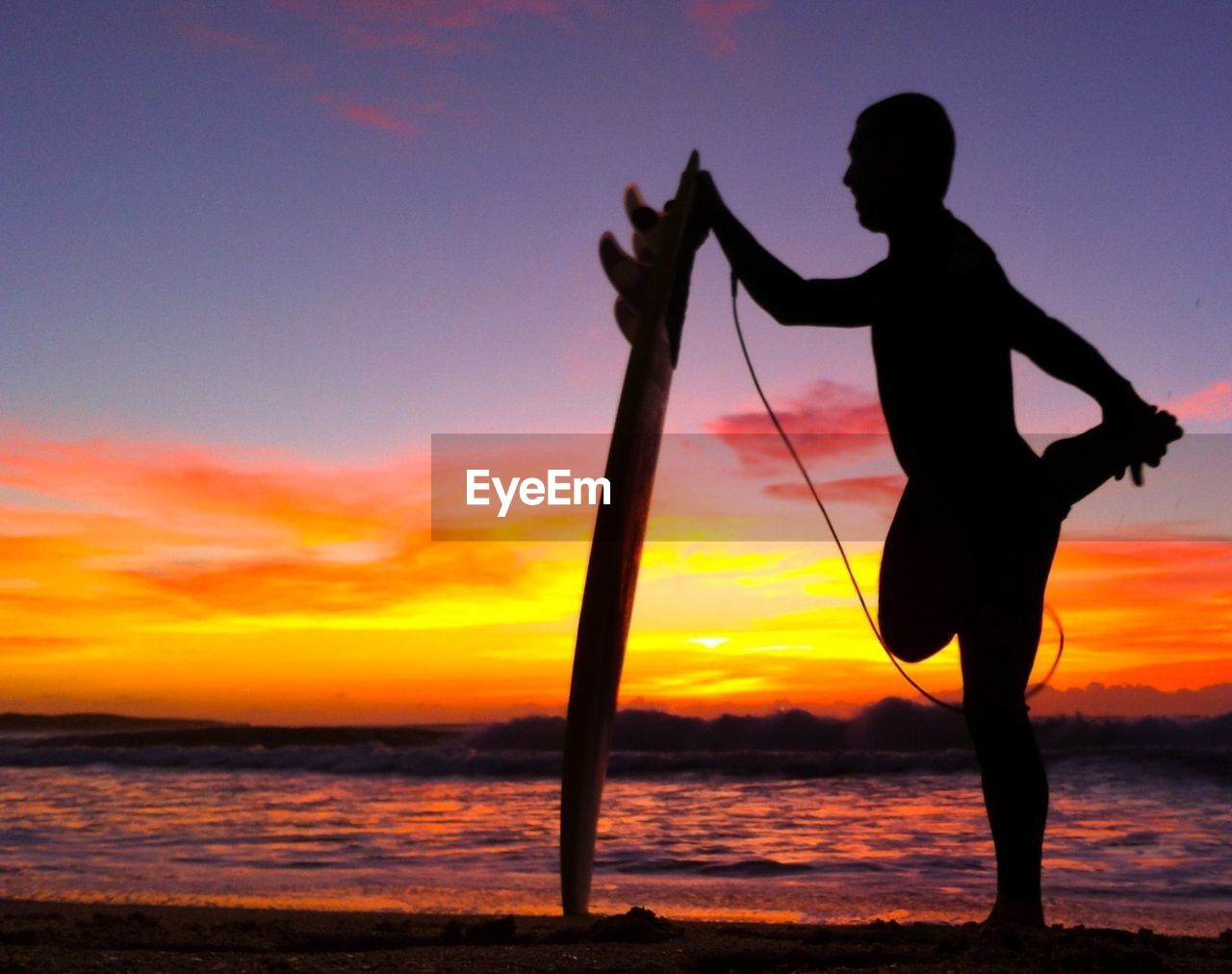 Silhouette man holding surfboard while stretching leg on beach at sunset