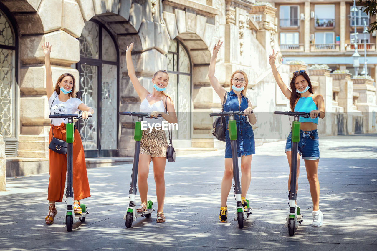 Young tourists with face masks having fun riding electric scooters while exploring city