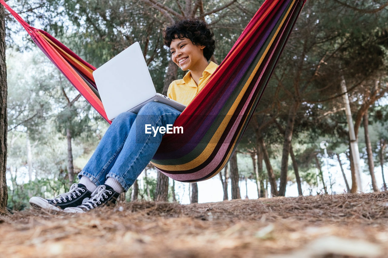Ground level of smiling teen female chilling in hammock in park and browsing laptop