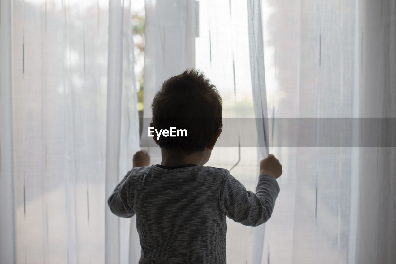 Rear view of boy standing by curtain at home