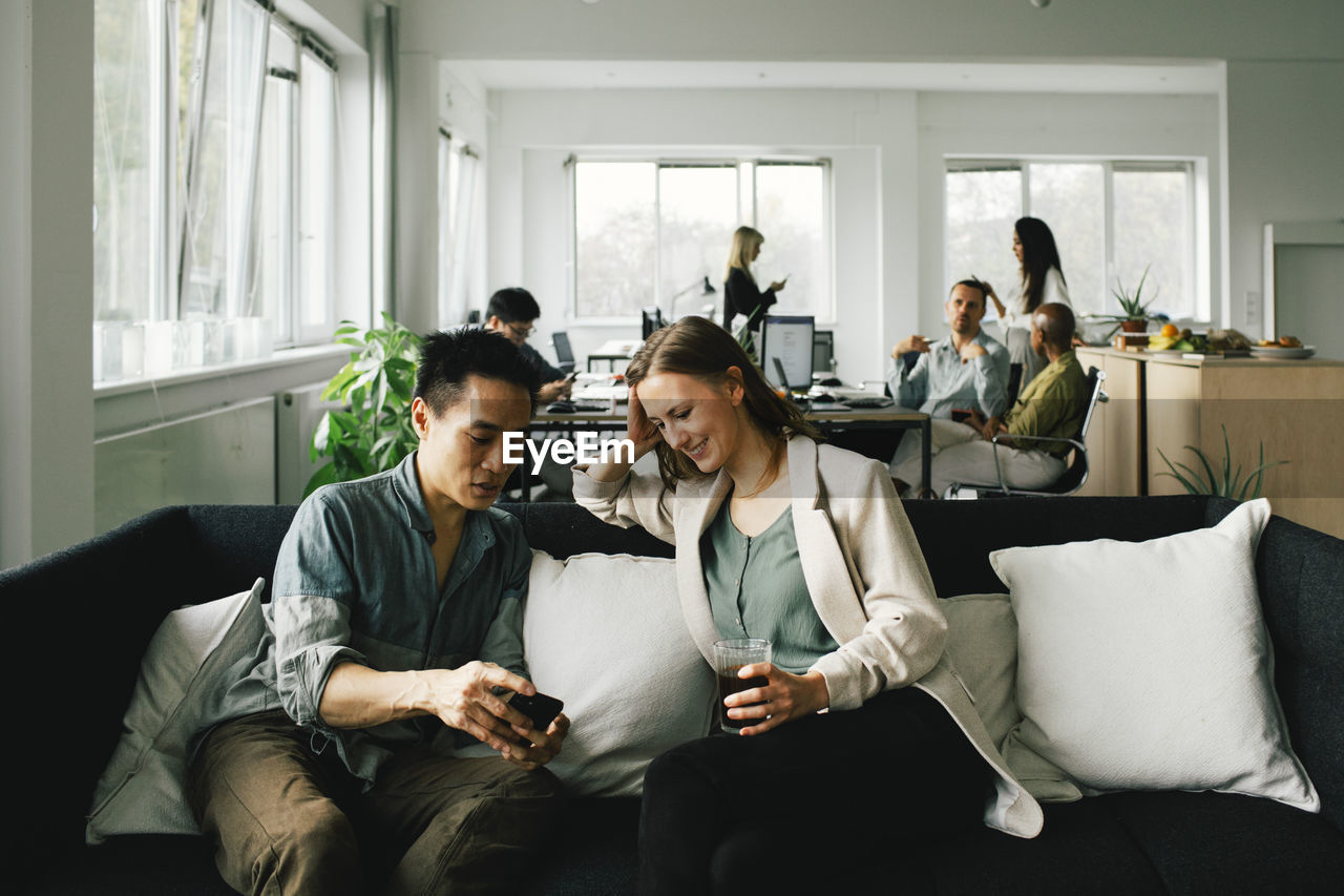 Businessman sharing smart phone while female colleague holding drink while sitting on sofa at home