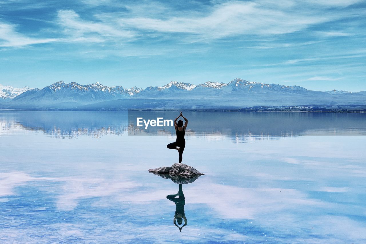 Woman doing yogapose on rock in lake against mountainrange