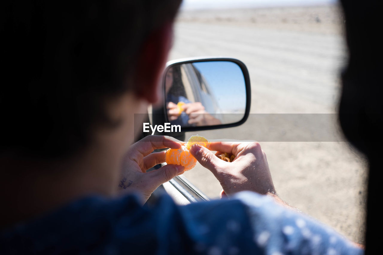 Cropped image of man peeling orange while sitting in car by reflection on side-view mirror