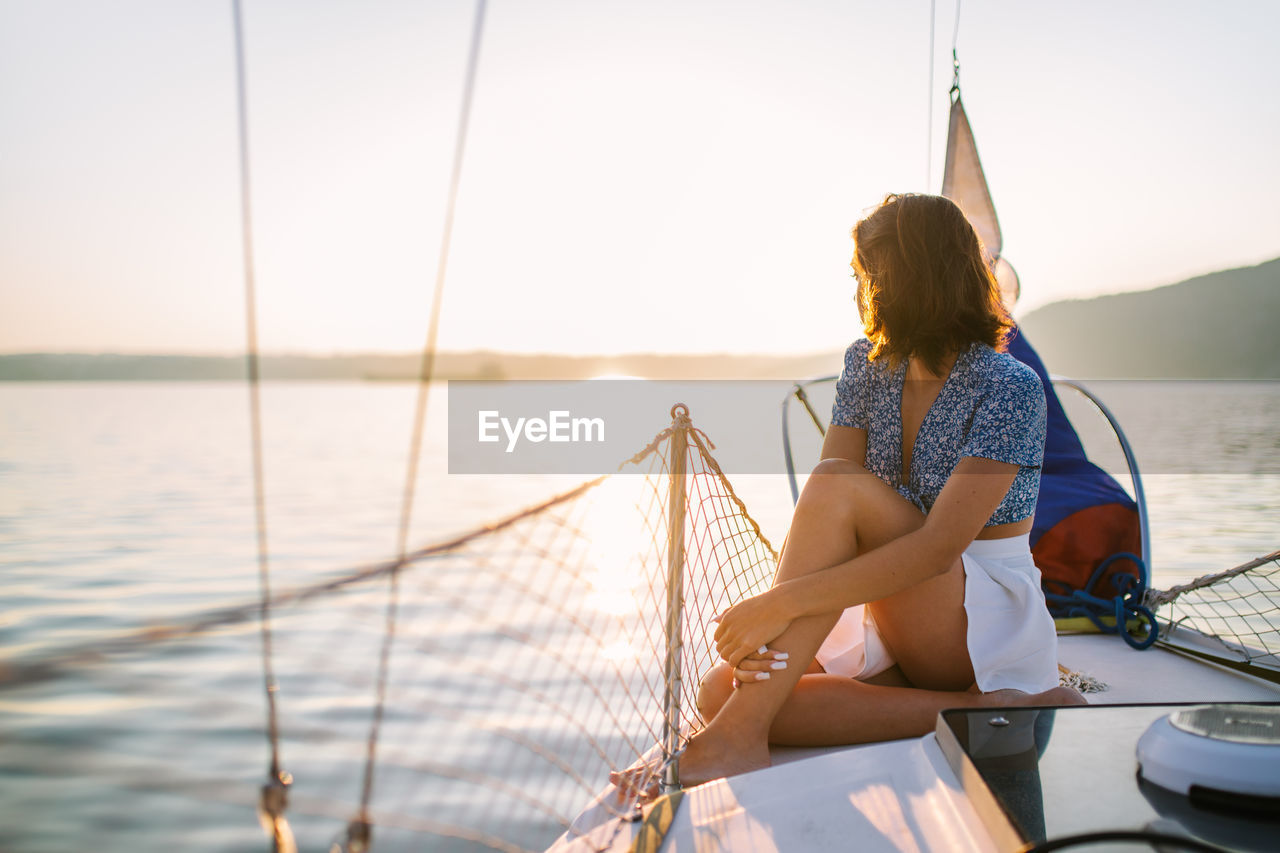 Side view of young stylish female traveler with dark hair in trendy crop top and skirt admiring sea while sitting on sailboat during cruise under sunset sky