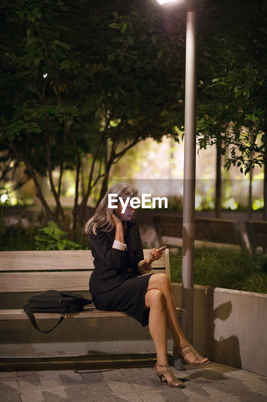 Full length of businesswoman using phone while sitting on bench at park