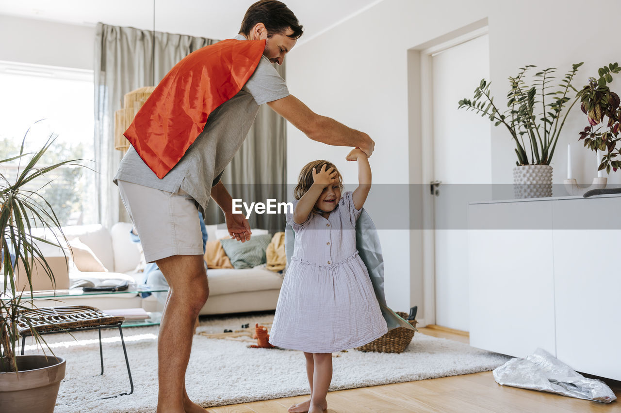 Side view of father wearing cape and dancing with daughter in living room at home