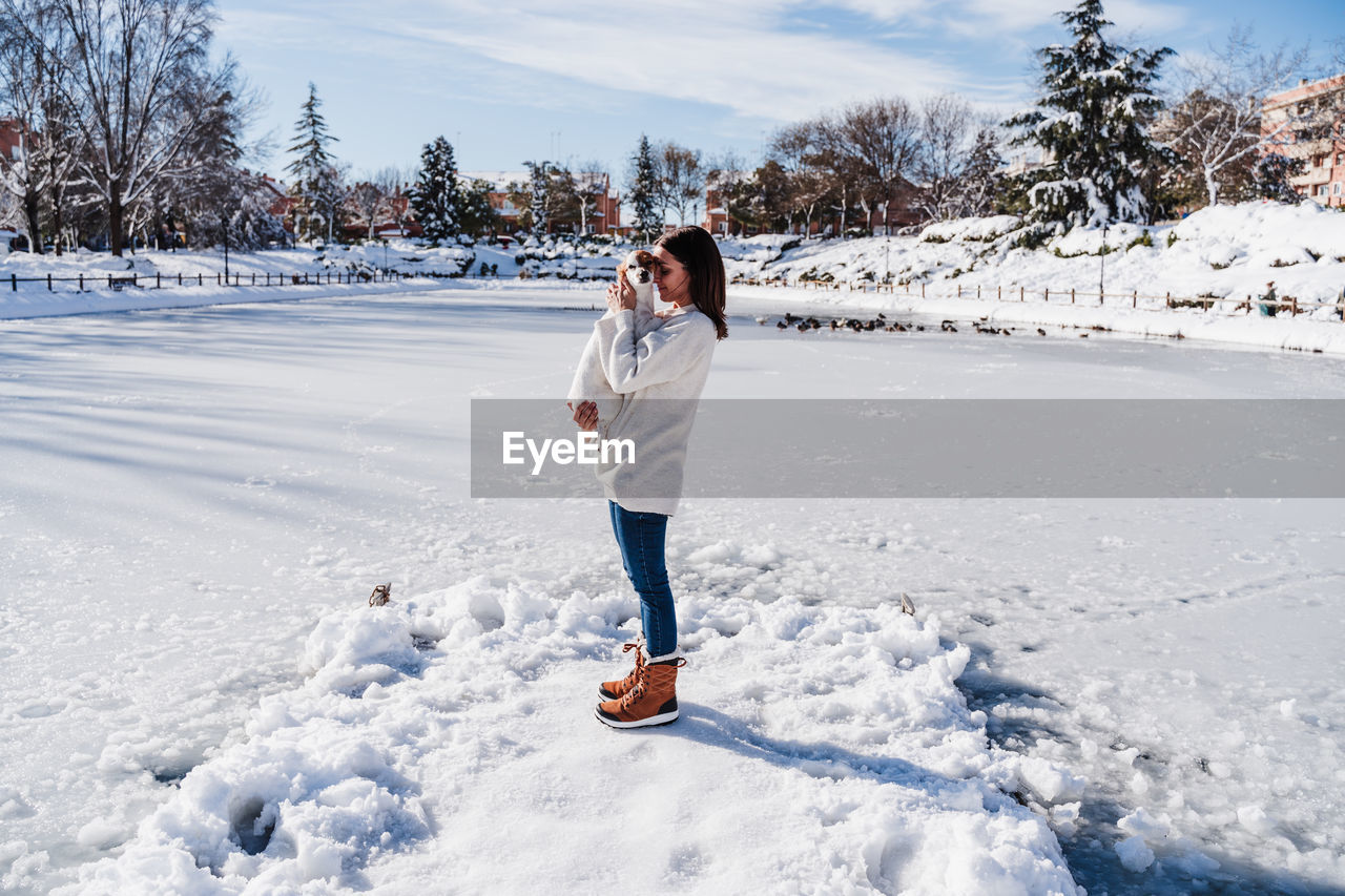 Woman hugging cute jack russell dog while standing on snowy pier by frozen lake during winter