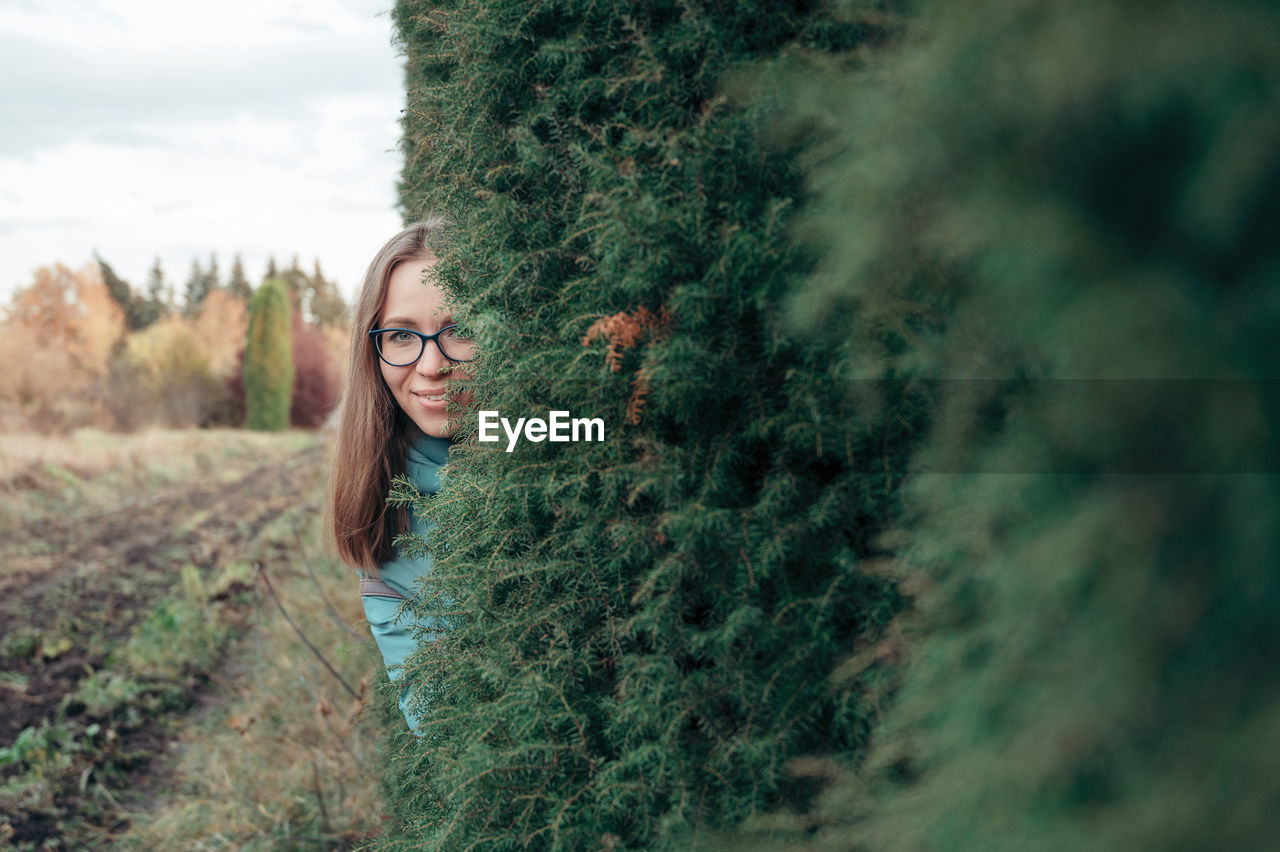 PORTRAIT OF YOUNG WOMAN STANDING BY PLANTS