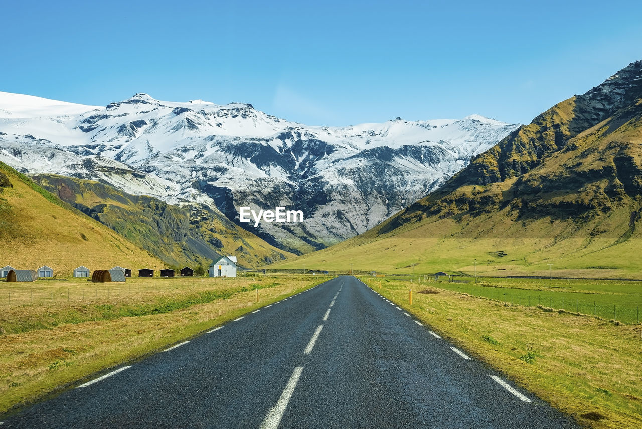 Diminishing empty road leading towards snowcapped mountain against clear blue sky