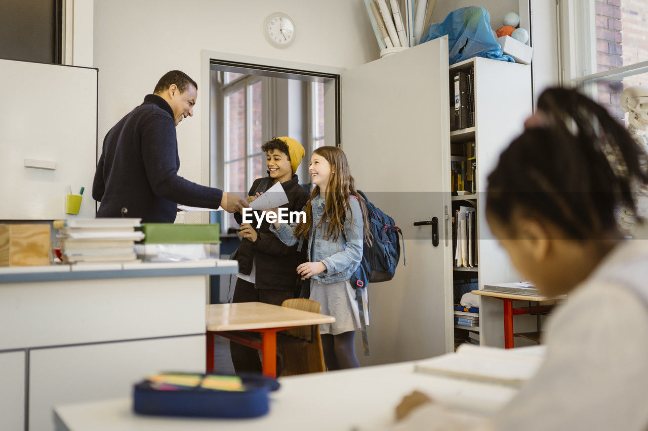 Mature male teacher giving documents to happy students standing in classroom