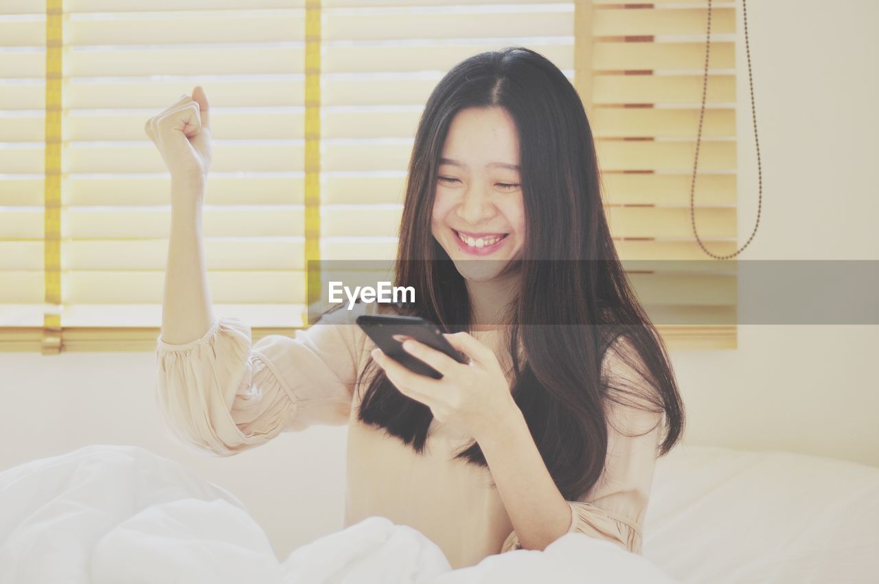 Happy young woman using smart phone on bed at home