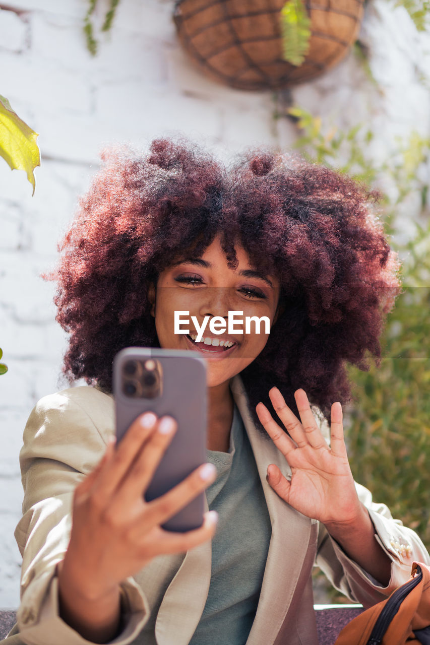 Cheerful african american female manager smiling and waving hand while making video call via cellphone on street